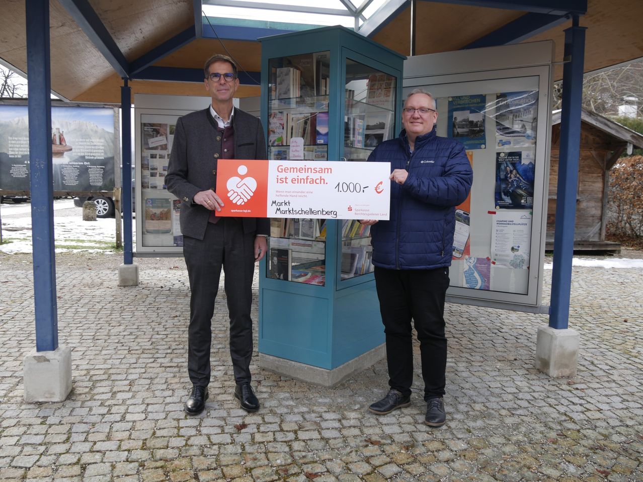 Zwei Männer stehen vor einem Bücherkiosk und halten einen Scheck über 1.000 Euro. Sie lächeln und scheinen für ein Foto zu posieren.