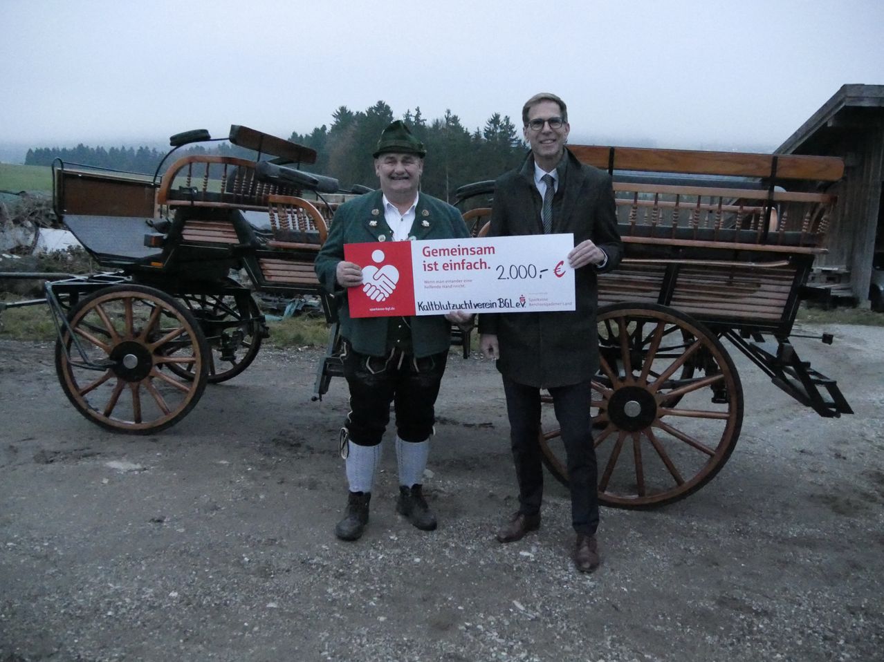 Two men stand in front of a carriage, one holding a large cheque for 2,000 euros. The man in traditional clothing smiles, and the other wears a formal suit.