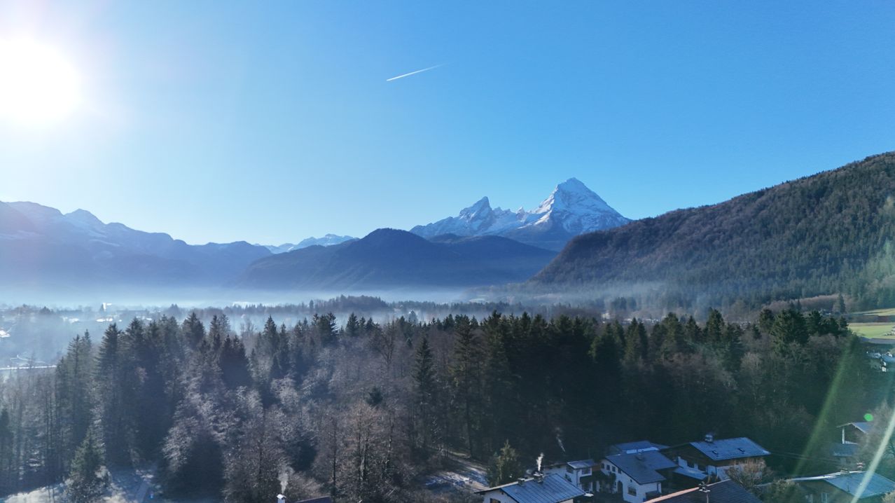 A high-altitude view shows a clear blue sky with a trail of a plane, snowy mountains in the background, and a valley with dense trees and fog.