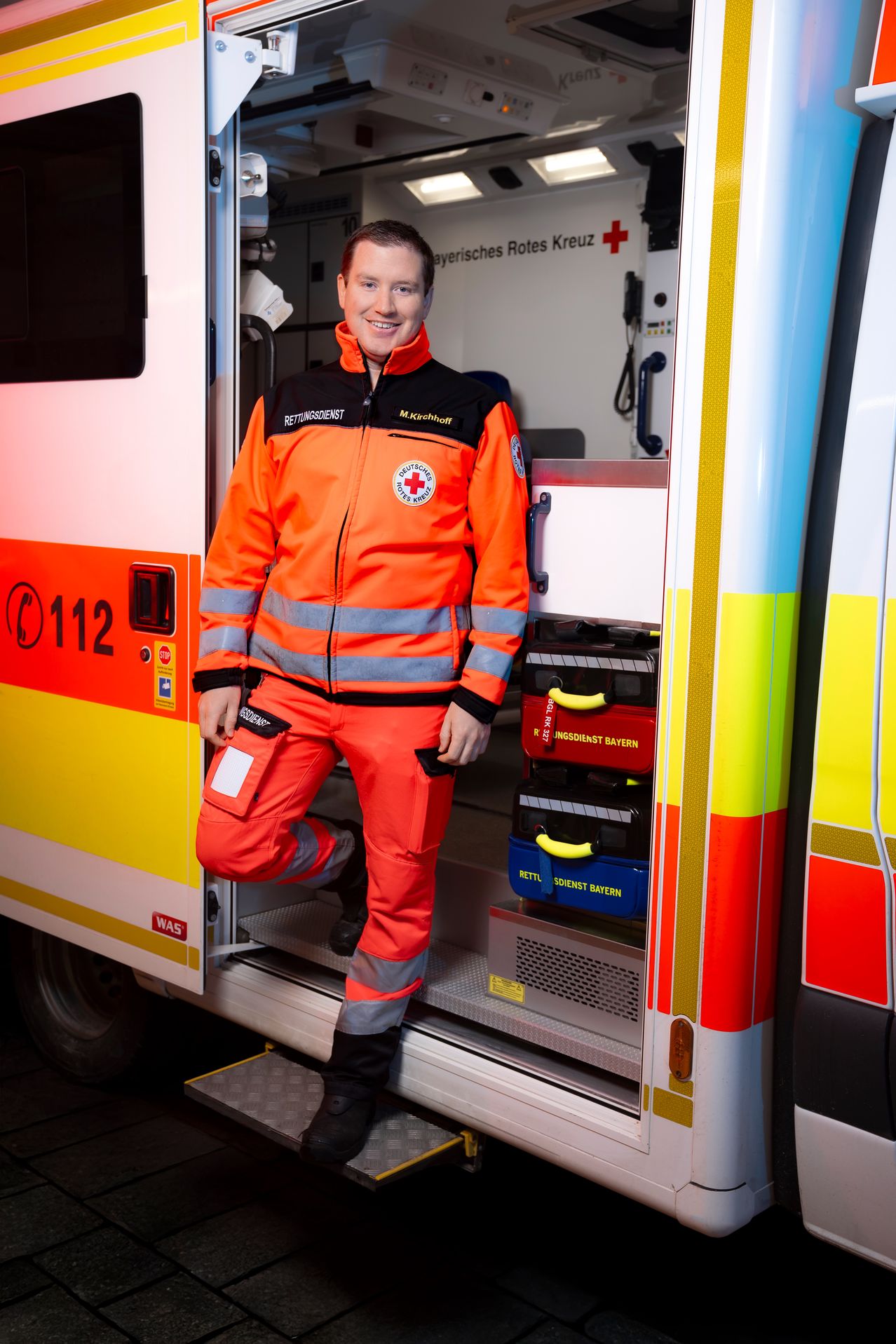Ein Rettungssanitäter mit Lächeln, gekleidet in einem orangefarbenen Uniform, steht neben einem Krankenwagen. Der Krankenwagen hat medizinische Ausrüstung im Inneren und die Nummer 112 an seiner Seite.