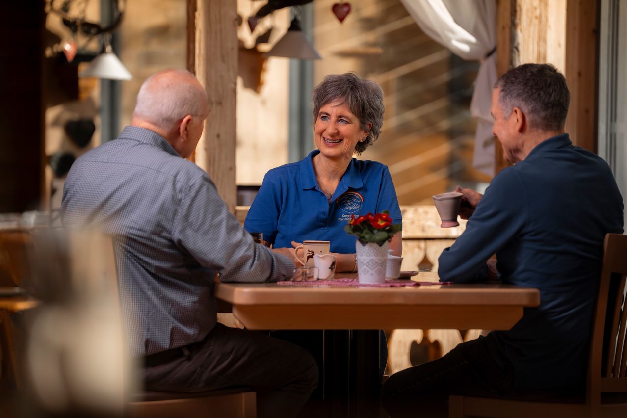 Three people sit around a table in a warm room. One woman smiles and the others look at her. There is a vase of flowers and cups on the table.