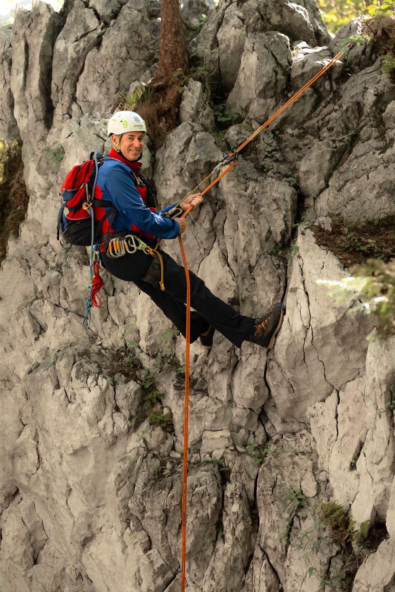 Eine Person klettert eine felsige und steile Felswand mit Seilen und Ausrüstung. Sie trägt einen Helm und einen Rucksack.