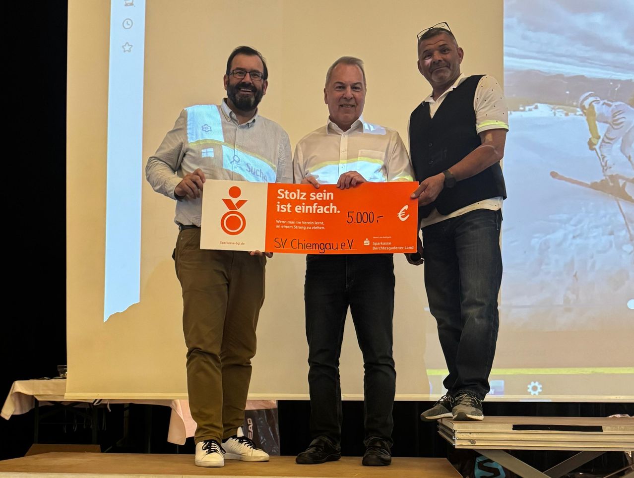 Drei Männer stehen auf einer Bühne und halten einen großen orangefarbenen Scheck über 5000 Euro. Sie lächeln und schauen in die Kamera. Der Text auf dem Scheck lautet Stolz sein ist einfach. Der Hintergrund zeigt eine Leinwand.