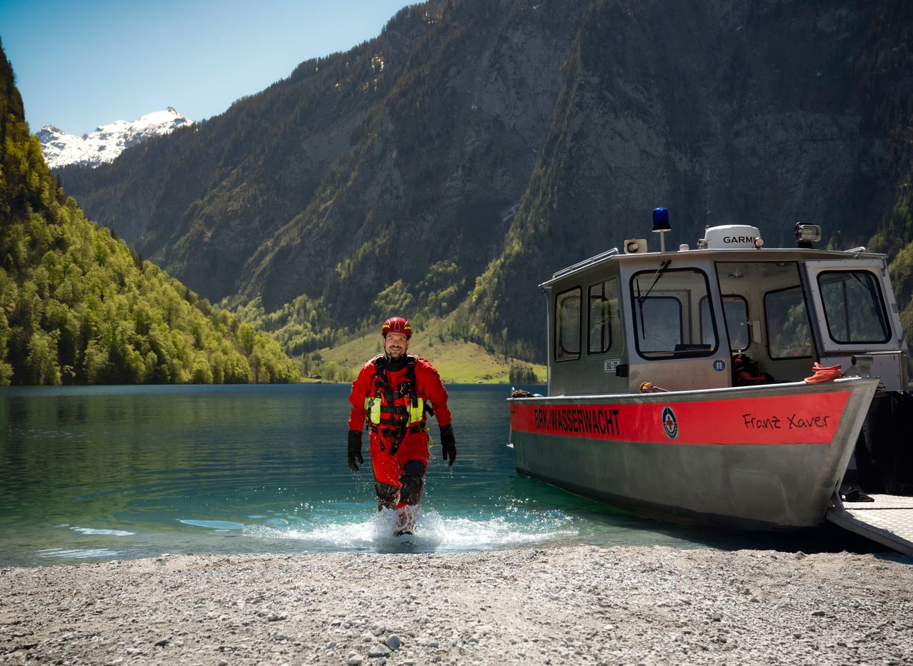 A man in a red suit walks on water near a boat with 'Front Wasserwacht' written on it. Mountains and a lake are in the background.