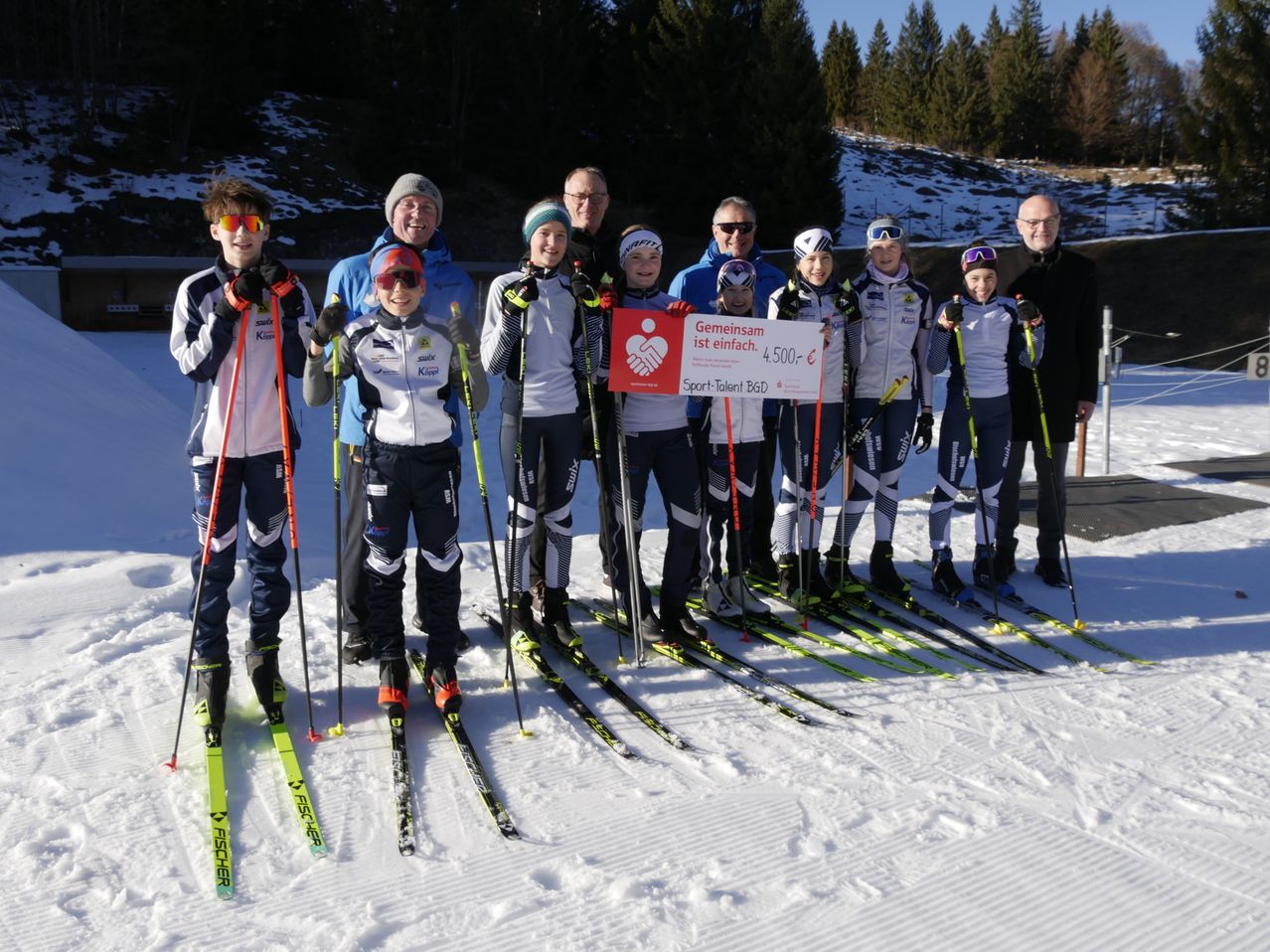 A group of skiers stand on snowy ground, holding ski poles. They pose with a check and a banner. Behind them, snowy hills and trees are visible.