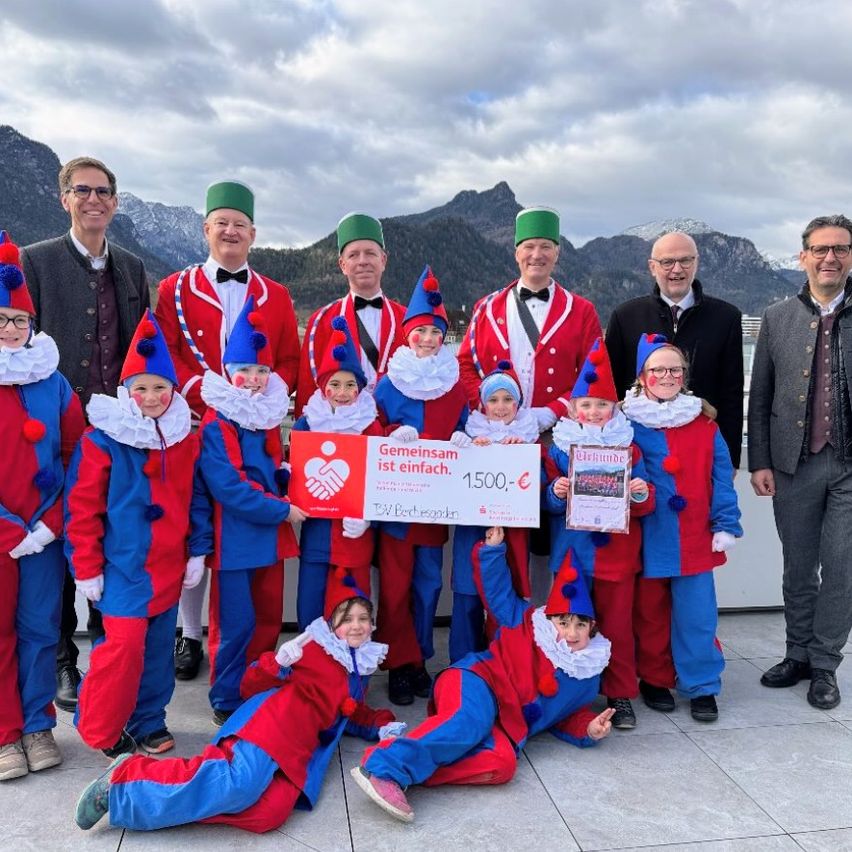 A group of children and adults dressed in clown costumes are standing together with some adults in front of a mountain range, holding a check for 1500 Euros.