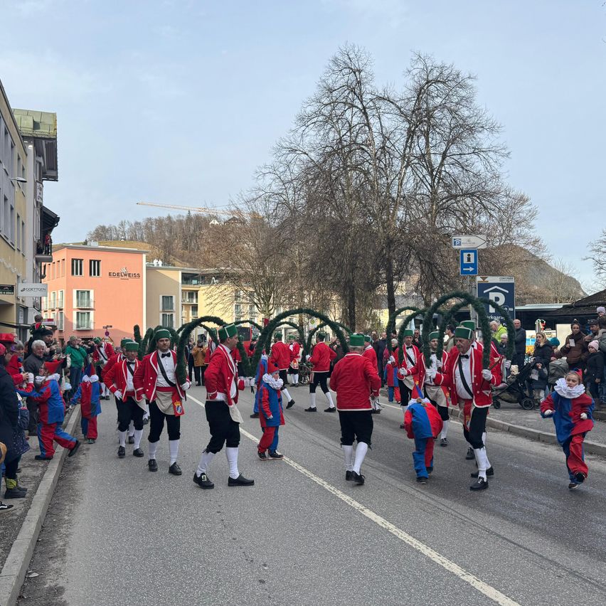 A street parade with participants dressed in red and green costumes marching, with onlookers on the sidewalk. Trees and buildings are visible along the street.