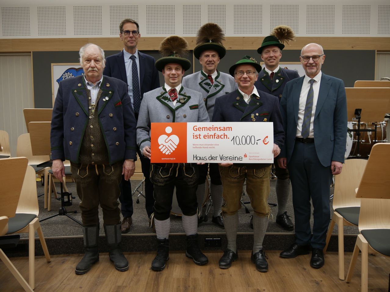 Eine Gruppe von Männern in traditioneller Kleidung posiert mit einem großen Scheck, stehend auf einer Bühne mit einem Holzboden und einer Kulisse. Der Scheck lautet 'Gemeinsam ist einfach. 10.000€'.