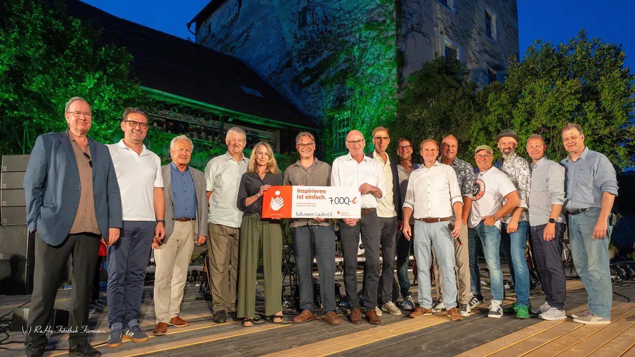 Eine Gruppe von Menschen steht auf einer Holzterrasse und hält einen großen Scheck über 7.000 Euro, mit einem Gebäude und grünen Lichtern im Hintergrund.