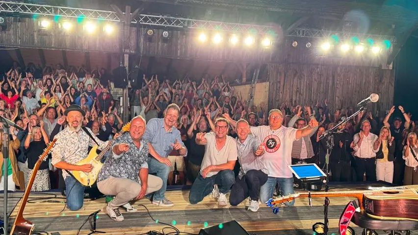Eine Band posiert mit Gitarren auf der Bühne und lächelt das Publikum an, das klatscht und jubelt. Die Bühne ist gut beleuchtet mit Scheinwerfern und einem Hintergrund aus Holzpaneelen.