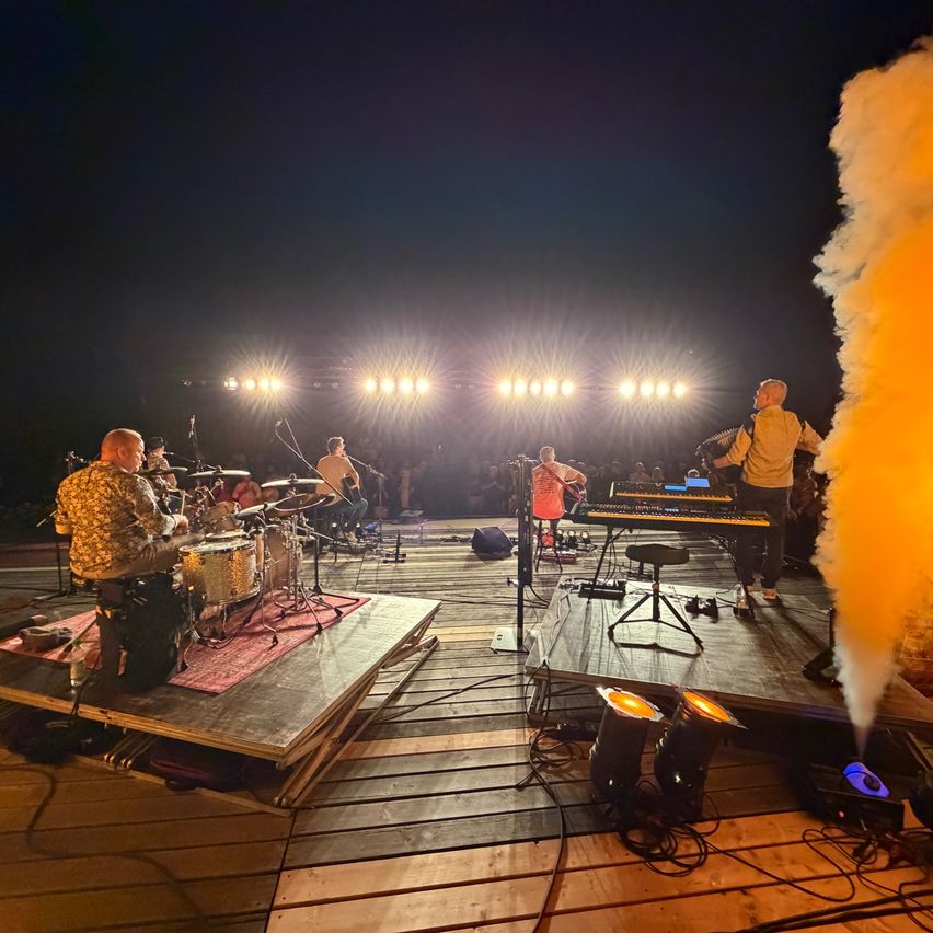 Eine Band spielt auf einer Holzbühne bei Nacht mit gelben Raucheffekten, bestehend aus einem Schlagzeuger, einem Keyboarder und einem Sänger.