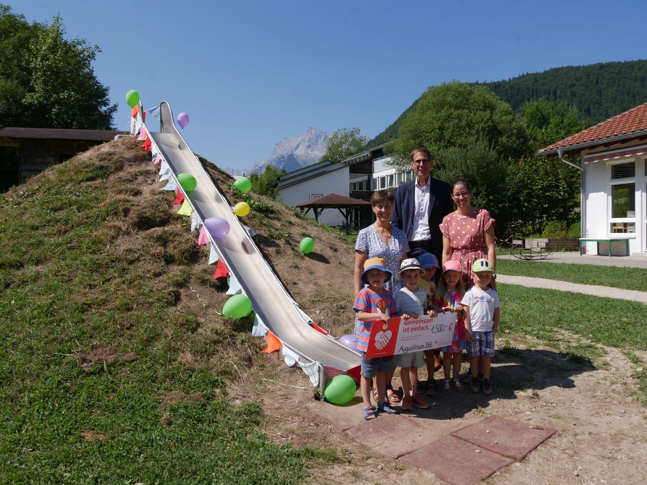 Eine Gruppe von Menschen, einschließlich Erwachsener und Kindern, steht vor einer großen Rutsche, die mit bunten Luftballons und Flaggen geschmückt ist. Sie halten einen großen Scheck und posieren für ein Foto. Hinter ihnen befinden sich Häuser und Berge im Hintergrund.