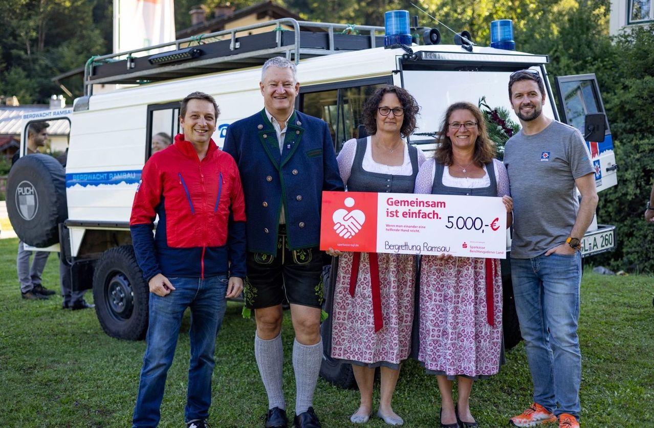 Fünf Personen stehen vor einem weißen Fahrzeug mit einem roten Scheck über 5.000 Euro. Sie lächeln und scheinen für ein Foto zu posieren.