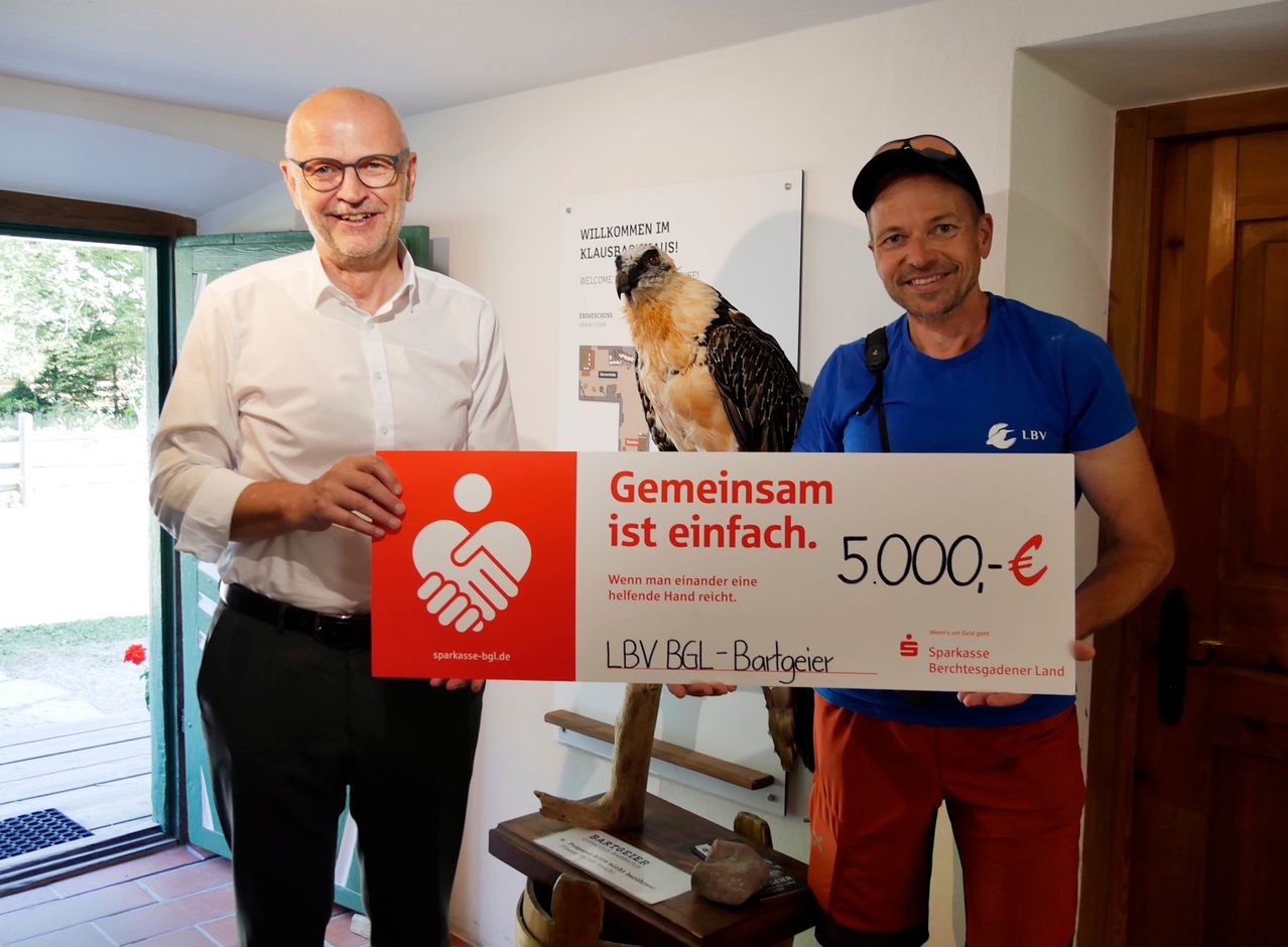 Two men hold a large sign with a bird on it. The sign says 'Together is simple.' The man on the left wears a white shirt and black pants. The man on the right wears a blue shirt and red shorts.
