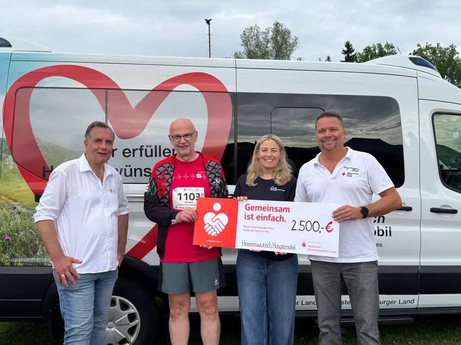 Vier Personen stehen vor einem Van mit einem Spendenscheck, einer hält ihn. Der Mann in Rot trägt eine Brille und ein Startnummernband mit der Nummer 1103.