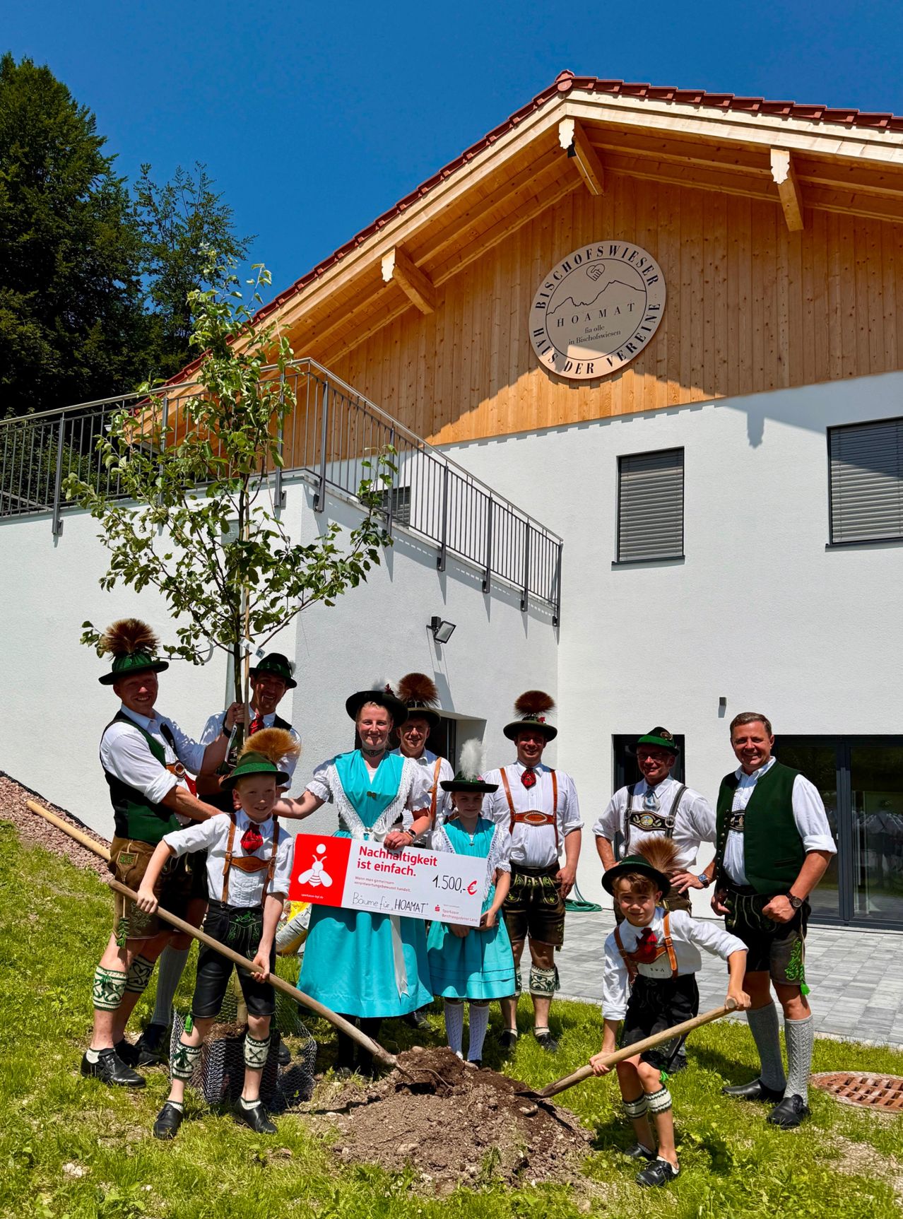 Eine Gruppe von Menschen in traditioneller Kleidung pflanzt einen Baum vor einem Gebäude. Sie halten ein Zertifikat und Schaufeln. Das Gebäude hat ein Schild, das Bischofswiesen Heimat besagt.