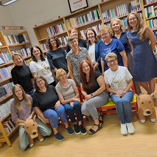 Eine Gruppe von Frauen und Mädchen posiert für ein Foto in einer Bibliothek mit Bücherregalen im Hintergrund. Einige sitzen auf einer Bank, während andere stehen.