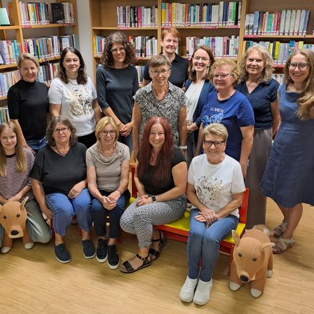 Eine Gruppe von Frauen in lässiger Kleidung, sitzend und stehend, posiert für ein Foto in einer mit Bücherregalen gefüllten Bibliothek. Einige tragen Brillen, und zwei haben Hundespielzeug auf dem Schoß.