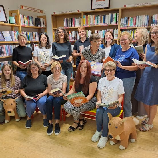 Eine Gruppe von Frauen, einige sitzend und andere stehend, posiert für ein Foto in einer Bibliothek, alle halten Bücher und lächeln.