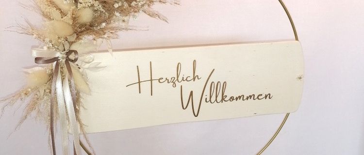 Ein runder Kranz mit getrockneten Blumen und einem weißen Schild mit den Worten Herzlich Willkommen. Der Kranz ist mit einer Schleife verziert und auf einem weißen Hintergrund platziert.
