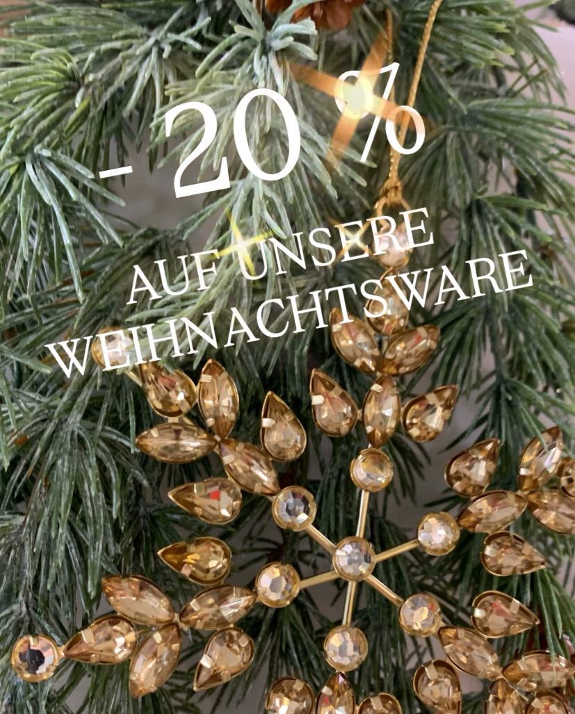 Ein goldener Schneeflocken-Schmuck hängt an einem Weihnachtsbaum. Der Schmuck hat den Text '-20% AUF UNSERE WEIHNACHTSWARE' darauf. Der Baum ist mit grünen Blättern dekoriert.