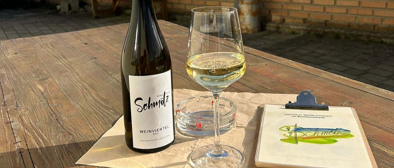 Eine Flasche Wein und ein Glas stehen auf einem Holztisch, neben einer Speisekarte und einem Klemmbrett, vor dem Hintergrund einer Backsteinmauer und eines Gebäudes.