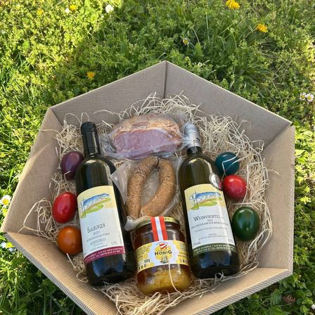 Eine Pappschachtel enthält zwei Weinflaschen, ein Glas Honig, eine Wurst, ein Stück Schinken und drei Ostereier auf einem Grasfeld.