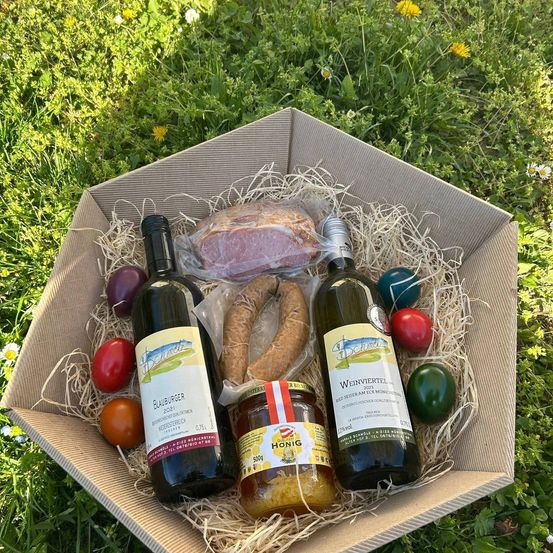 Eine Pappschachtel enthält zwei Weinflaschen, ein Glas Honig, eine Wurst, ein Stück Schinken und drei Ostereier auf einem Grasfeld.