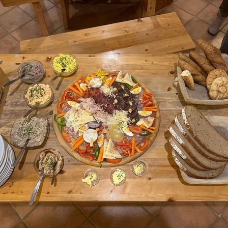 Ein Holztisch mit verschiedenen Gerichten, darunter Eier, Fleisch, Gemüse und Brot auf Tellern und Schüsseln. Eine Holzbank ist ebenfalls sichtbar.
