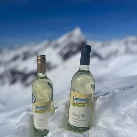 Zwei Weinflaschen, eine mit dem Aufkleber Geischter Satz 2022, sind im Schnee vergraben, mit einem Gebirgszug im Hintergrund.