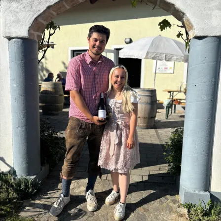 Ein Mann und eine Frau stehen lächelnd in einem Weinberg. Sie halten eine Flasche Wein. Hinter ihnen stehen Fässer und ein Schirm. Es gibt einen Tisch und Bänke.