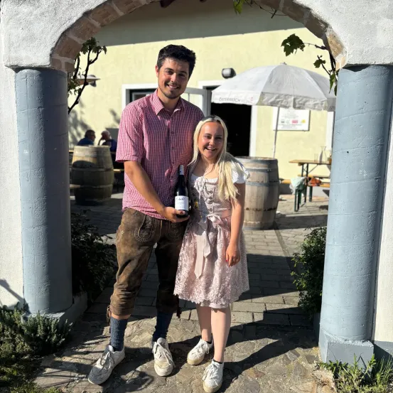 Ein Mann und eine Frau stehen lächelnd in einem Weinberg. Sie halten eine Flasche Wein. Hinter ihnen stehen Fässer und ein Schirm. Es gibt einen Tisch und Bänke.