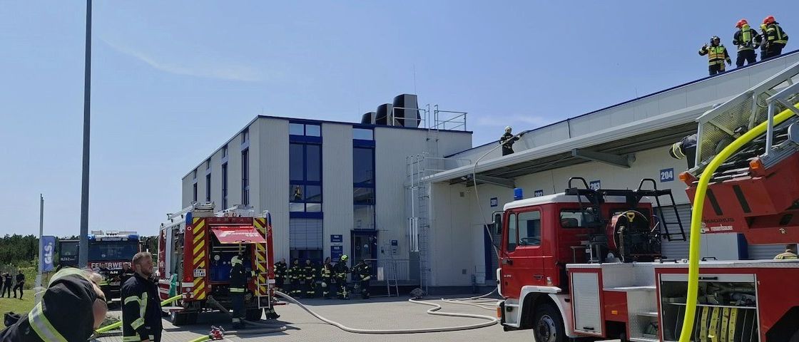 Feuerwehrleute sind bei einem Gebäude mit mehreren Feuerwehrwagen. Ein Feuerwehrmann ist auf dem Dach. Es sind Feuerwehrschläuche an die Wagen angeschlossen.