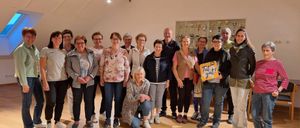 Eine Gruppe Erwachsener posiert für ein Foto in einem Raum mit Holzböden. Einige tragen Brillen und Jacken. Eine hält ein Schild mit der Aufschrift 'Tut gut!'. Trophäen sind an der Wand ausgestellt.