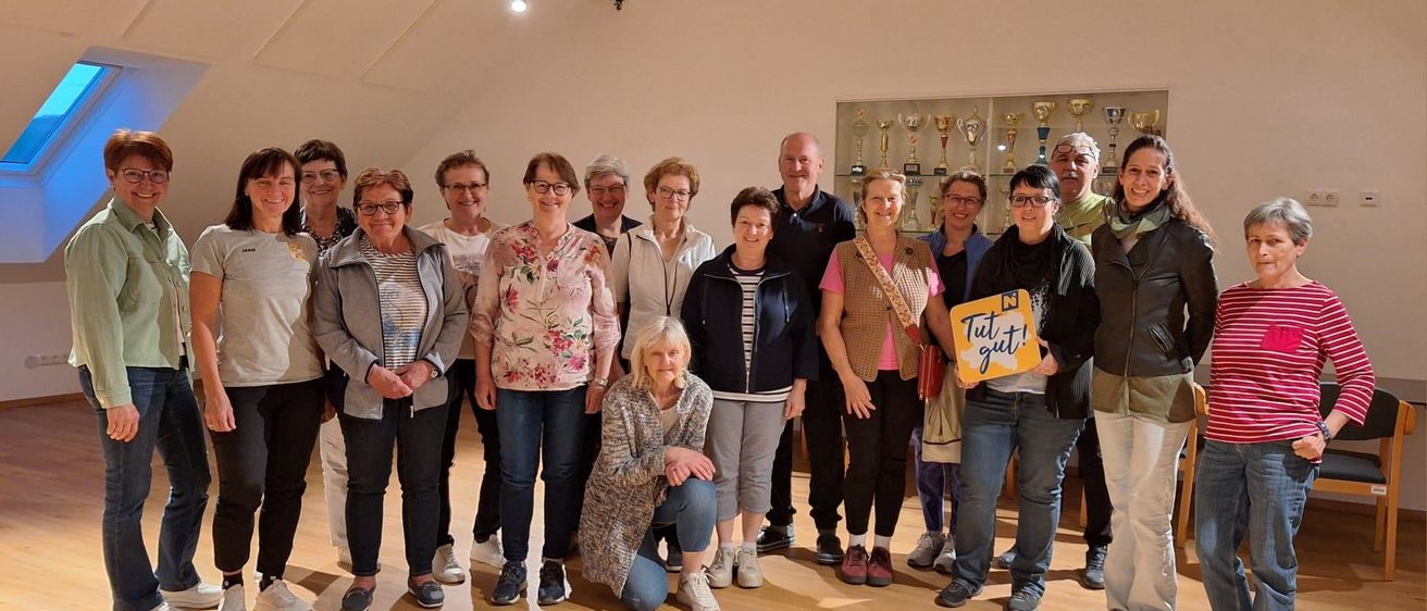 Eine Gruppe Erwachsener posiert für ein Foto in einem Raum mit Holzböden. Einige tragen Brillen und Jacken. Eine hält ein Schild mit der Aufschrift 'Tut gut!'. Trophäen sind an der Wand ausgestellt.