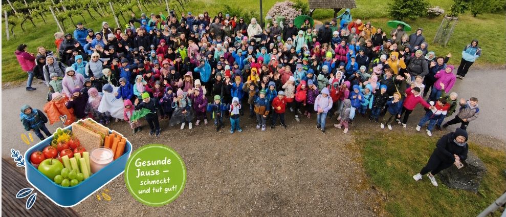 Eine große Gruppe von Kindern und Erwachsenen posiert für ein Foto auf einem Hügel. Im Hintergrund ist eine üppige Landschaft mit Hügeln und Bäumen zu sehen. Sie halten verpackte Mittagessen in den Händen.