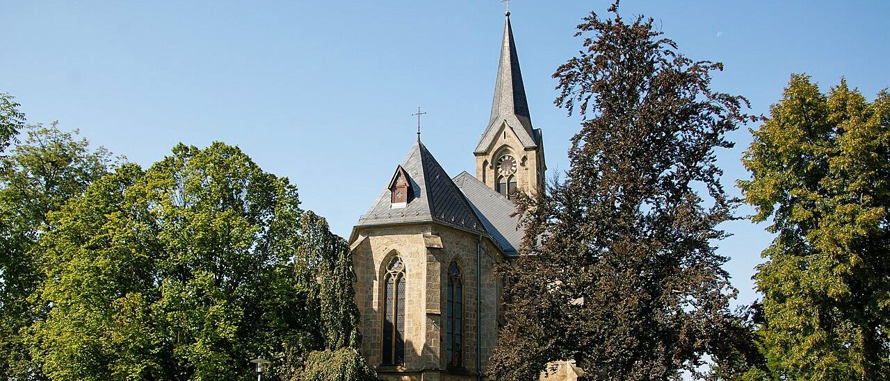 Eine alte Kirche mit einem spitzen Turm und einer Uhr steht vor einem hellblauen Himmel. Die Kirche ist von üppigem grünen Laub und Gras umgeben.