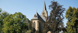 Eine alte Kirche mit einem spitzen Turm und einer Uhr steht vor einem hellblauen Himmel. Die Kirche ist von üppigem grünen Laub und Gras umgeben.