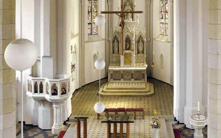 Ein Innenraum einer Kirche mit einem zentralen Altar, einem Kreuz und Buntglasfenstern. Der Altar ist von einer goldgeränderten Plattform umgeben. Drei Bänke sind in der Nähe platziert.