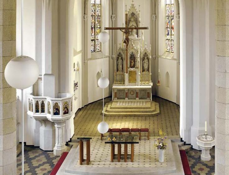 Ein Innenraum einer Kirche mit einem zentralen Altar, einem Kreuz und Buntglasfenstern. Der Altar ist von einer goldgeränderten Plattform umgeben. Drei Bänke sind in der Nähe platziert.