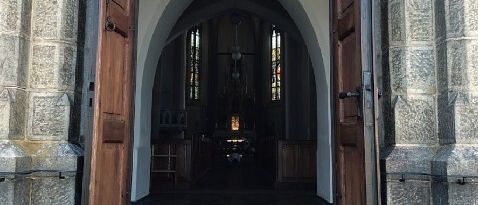 Der Eingang zu einer Kirche mit Holztüren und einem gefliesten Boden. Im Inneren gibt es einen Altar und Fenster.