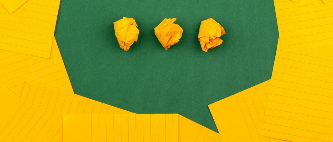Drei zerknitterte gelbe Papiere liegen auf einem grünen Hintergrund in einer Sprechblase aus gelbem Papier.