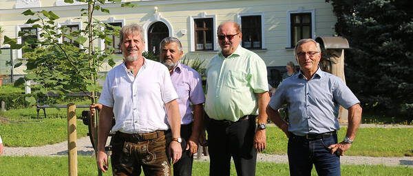 Vier Männer stehen in einem Park neben einer frisch gepflanzten Baum, in Freizeitkleidung. Hinter ihnen befindet sich ein großes, weißes Gebäude mit vielen Fenstern und einem roten Dach.