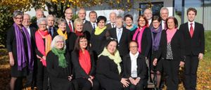 Eine Gruppe von Menschen in formeller Kleidung, einige sitzend und andere stehend, posiert für ein Foto vor einem Baum und einem Gebäude.