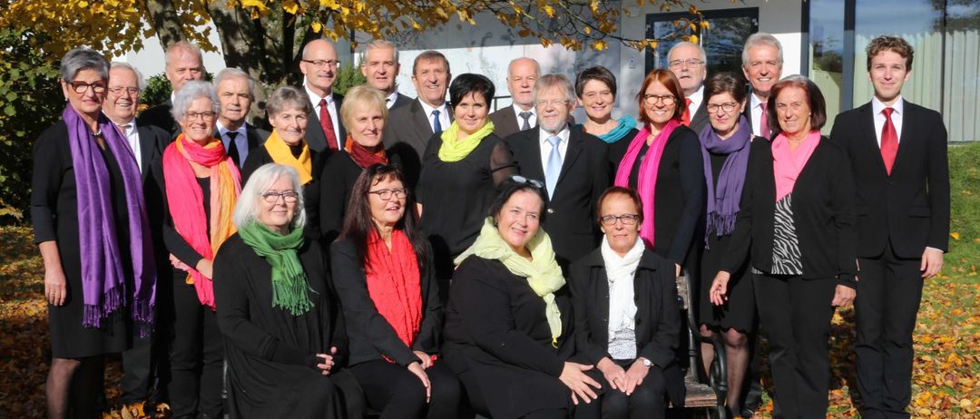 Eine Gruppe von Menschen in formeller Kleidung, einige sitzend und andere stehend, posiert für ein Foto vor einem Baum und einem Gebäude.