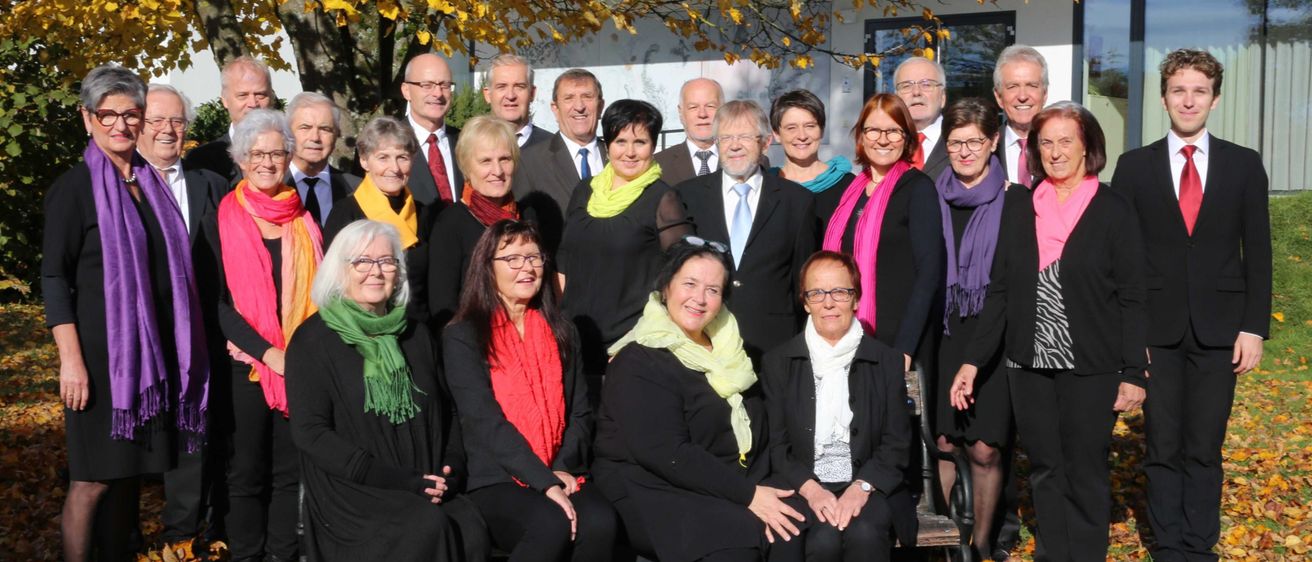 Eine Gruppe von Menschen in formeller Kleidung, einige sitzend und andere stehend, posiert für ein Foto vor einem Baum und einem Gebäude.