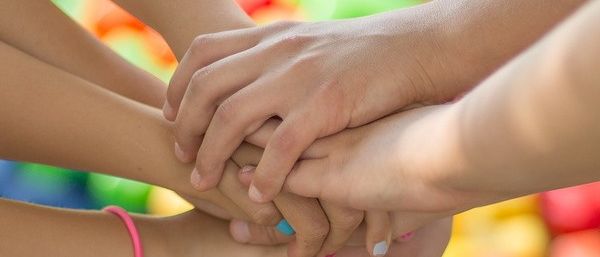 Mehrere Kinder mit bunten Armbändern an den Handgelenken halten in einer verspielten Anordnung Händchen. Dahinter sind bunte Bauklötze auf dem Boden verstreut.