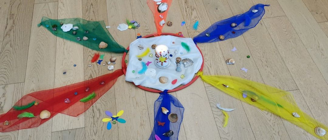 Eine kreisförmige Anordnung von bunten Federn, Muscheln und einer Kerze auf einem Holzboden, umgeben von mehrfarbigen Bändern.