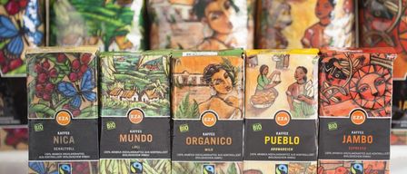 Mehrere Schachteln Bio-Kaffee sind aufgereiht mit Illustrationen von Menschen und Landschaften. Die Schachteln sind als Mundo, Organico und Pueblo gekennzeichnet. Sie sind Teil eines Fairtrade-zertifizierten Produkts.