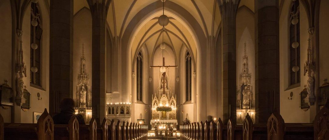 A dimly lit cathedral interior features a central aisle with glowing candles, a grand altar adorned with a cross, and statues of saints on the walls.